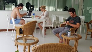 Ein Zimmer mit einem großen Fenster, weißen Stühlen, Korbmöbeln und einem Tisch mit Laptop, Wasserflasche und Tasse.