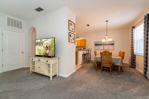 A dining area with a table and chairs, a television on a stand, and a kitchen in the background.