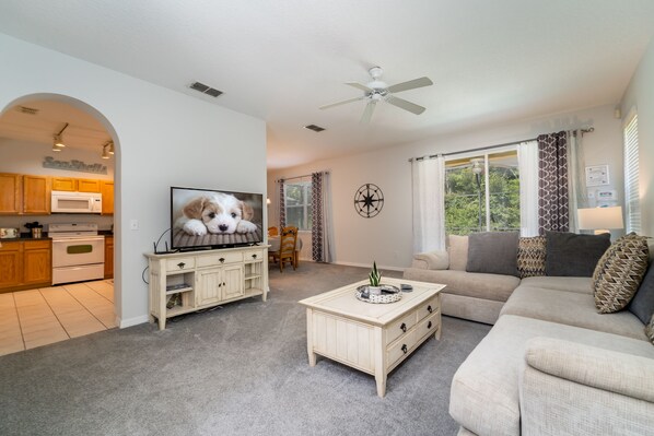 A living room with a TV, sofa, coffee table, and a kitchen area visible through an archway.