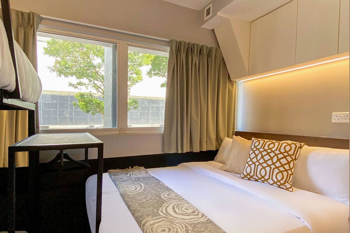 A hotel room with a bed, a desk, a window with curtains, and a headboard with built-in lighting.