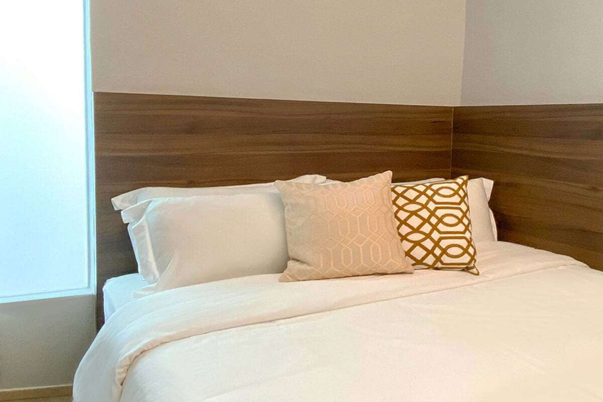 A neatly made bed with white bedding and two patterned pillows, a wooden headboard, and a window with curtains.