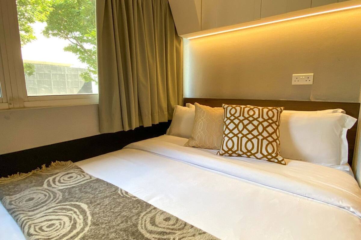 A hotel room with a bed, a window with curtains, a headboard with built-in lighting, and a power outlet on the wall.