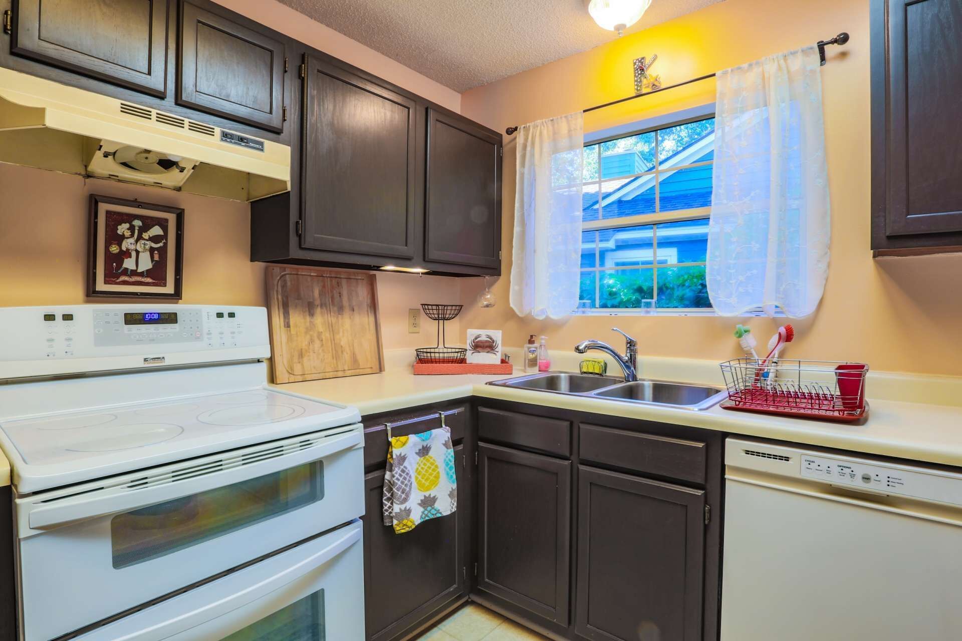 A kitchen with dark cabinets, a white stove, a sink, and a window with curtains.
