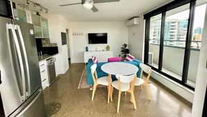 A compact kitchen with a refrigerator, microwave, and stove. There's a dining area with a table and chairs, a ceiling fan, and a large window offering a view of buildings.