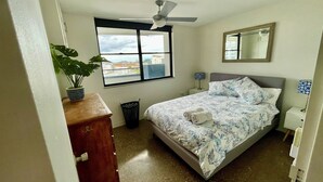 A bedroom with a bed, a ceiling fan, a window with a city view, a dresser, and a potted plant.