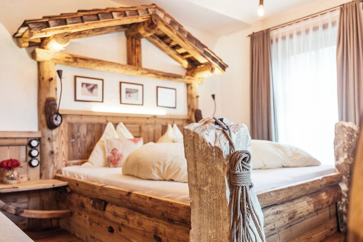 A rustic wooden bed with a sloped roof, a bedside table with a vase, and a window with curtains.