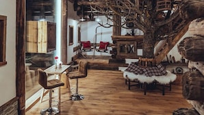 Un intérieur rustique avec un escalier en bois, un coin repas comprenant une table et des chaises, et un grand tronc d’arbre qui constitue l’élément central de la pièce.