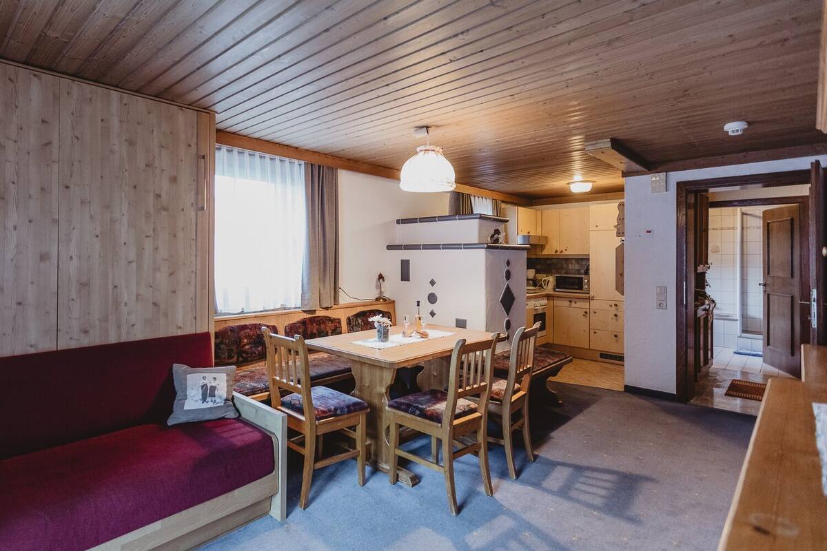 A cozy cabin interior with a wooden dining table, chairs, a red cushioned bench, and a kitchen area with a microwave and stove.
