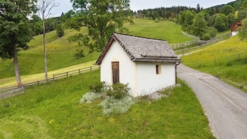 Ein kleines, weißes Gebäude mit brauner Tür, umgeben von grünem Gras und Bäumen, neben einer asphaltierten Straße.