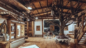 Un intérieur rustique avec des murs en bois, un grand tronc d’arbre servant de pièce maîtresse, un coin repas avec une table et des chaises, et une cheminée.