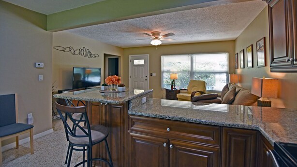 A kitchen with granite countertops, wooden cabinets, a ceiling fan, and a dining area with a TV and chairs.