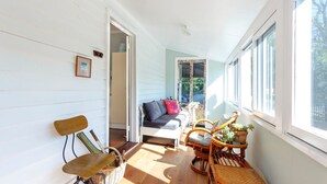 Une pièce chaleureuse avec un plancher en bois, un canapé blanc agrémenté de coussins, un fauteuil en osier, une petite table avec une plante et une fenêtre donnant sur l’extérieur.