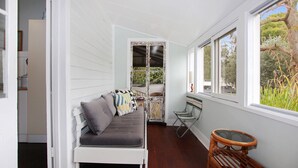 A narrow, enclosed porch with a white bench and cushions, a metal chair, and a glass table. - Sunroom Cottage - 40A Witt Street (Tea Gardens)