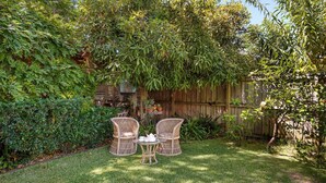 A garden with two wicker chairs and a small table, surrounded by lush greenery and a wooden fence. - Sunroom Cottage - 40A Witt Street (Tea Gardens)