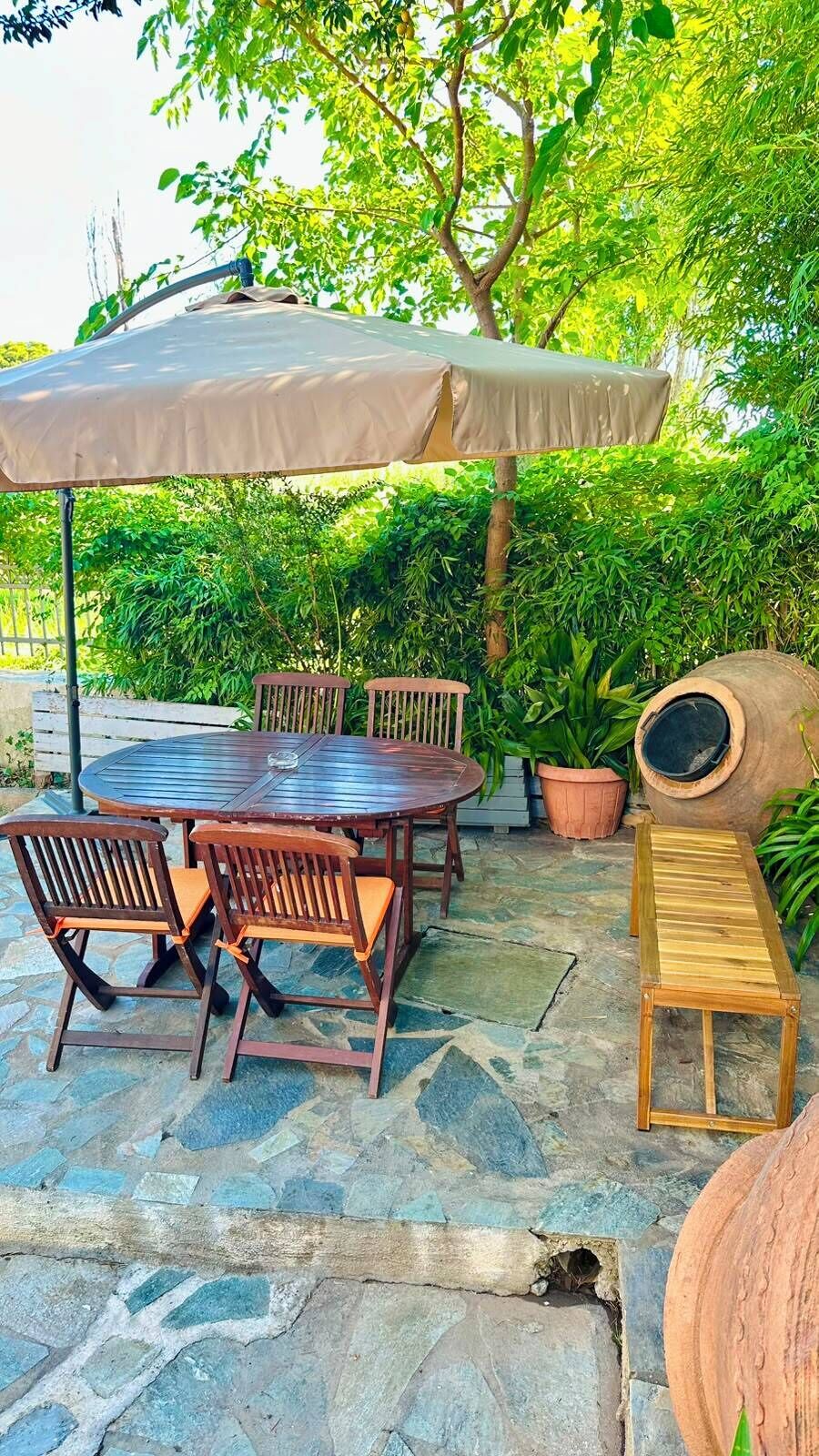 A round wooden table with chairs and a bench, an umbrella, and potted plants in an outdoor setting.