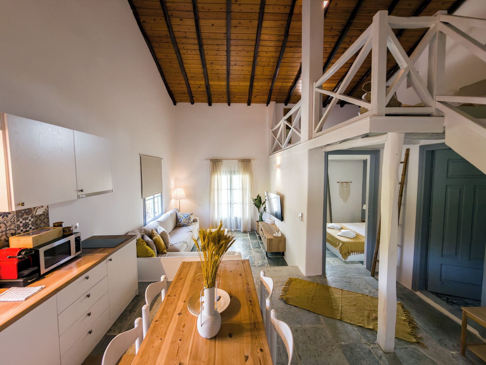 A spacious room with a wooden dining table, white chairs, a kitchen area with a microwave, and a staircase leading to another level.