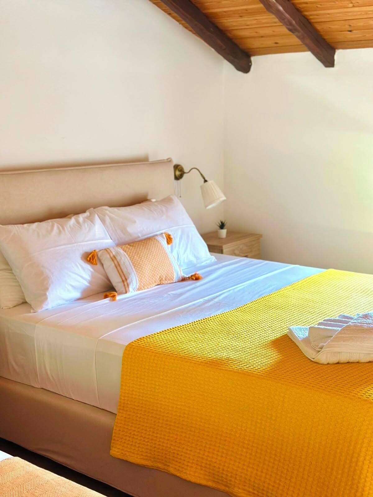 A bed with white bedding and a yellow throw blanket, a bedside lamp, and a small potted plant on a wooden nightstand.
