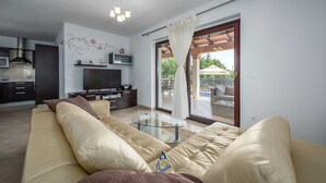 A modern living room with a beige sofa, a glass coffee table, and a flat-screen TV. There is a kitchen area with dark cabinets and a stainless steel range hood. A sliding glass door leads to an outdoor patio with a pool and umbrellas.