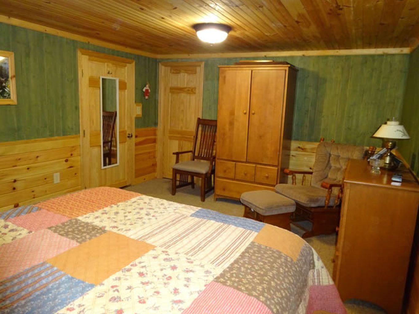 A bedroom with a large bed, a wooden wardrobe, a chair, and a desk with a lamp.