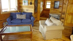A wooden cabin interior with a blue sofa, a beige armchair, and a glass-top coffee table.