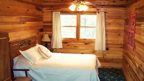 A wooden cabin bedroom with a bed, a chair, a nightstand, a lamp, and a window with curtains.