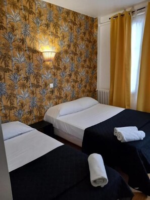 Une chambre d’hôtel avec deux lits, l’un recouvert d’une courtepointe blanche et l’autre d’une courtepointe noire. Une serviette enroulée est posée sur la courtepointe noire.