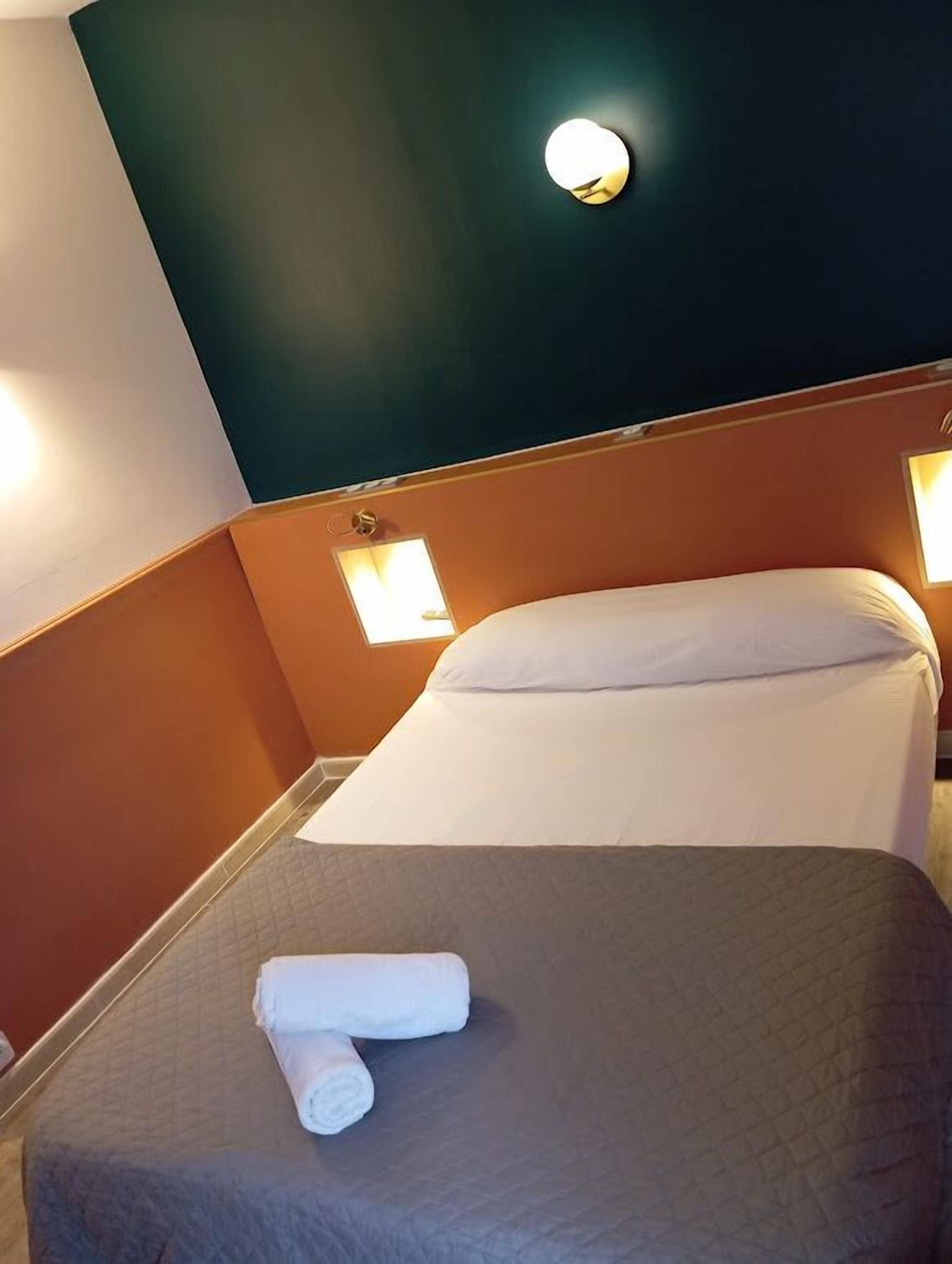 Un lit simple avec une couverture matelassée, une serviette enroulée et deux lampes fixées au mur.