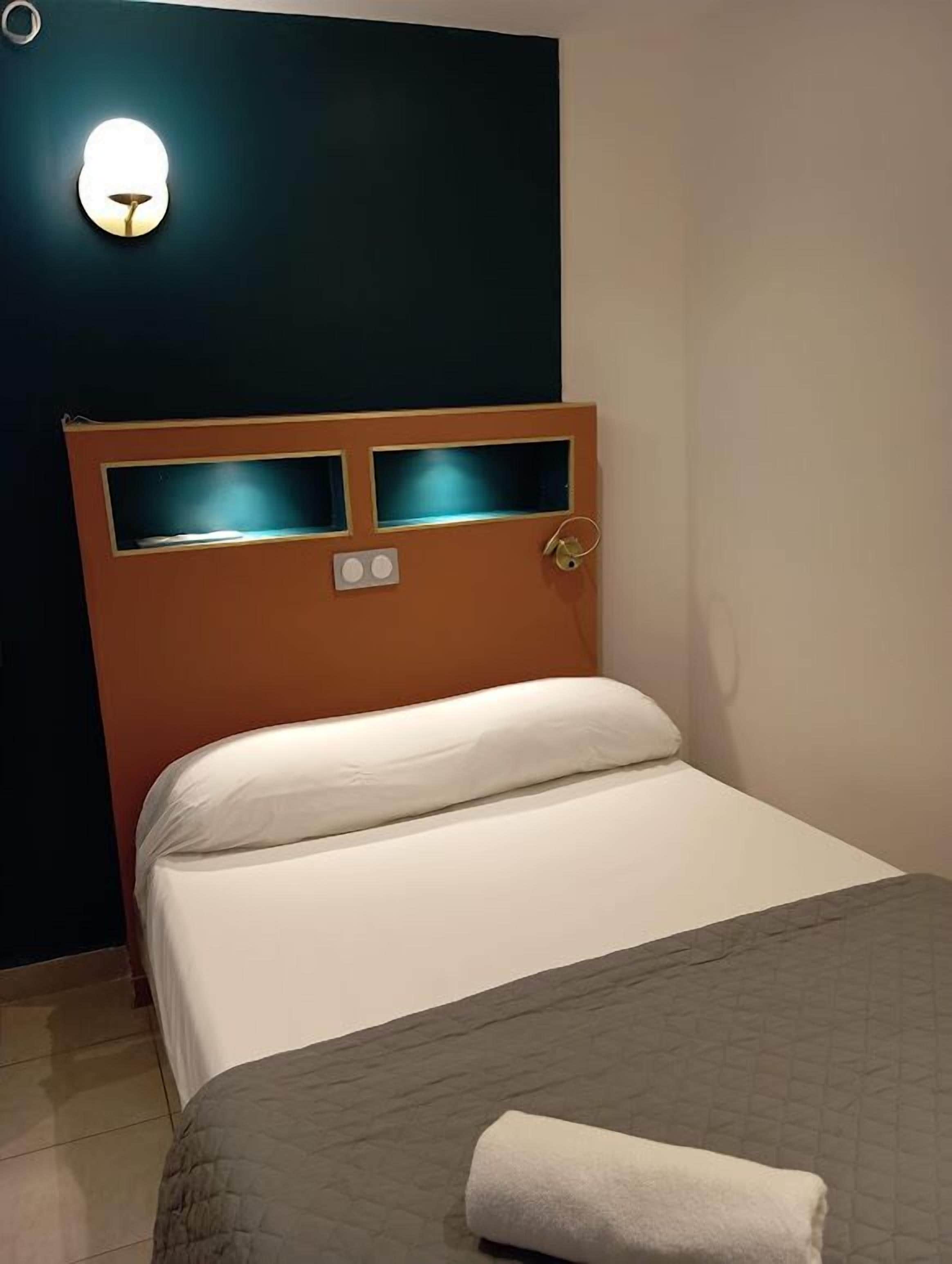 A hotel room with a bed, a headboard with two illuminated panels, a wall-mounted light, and a white pillow.