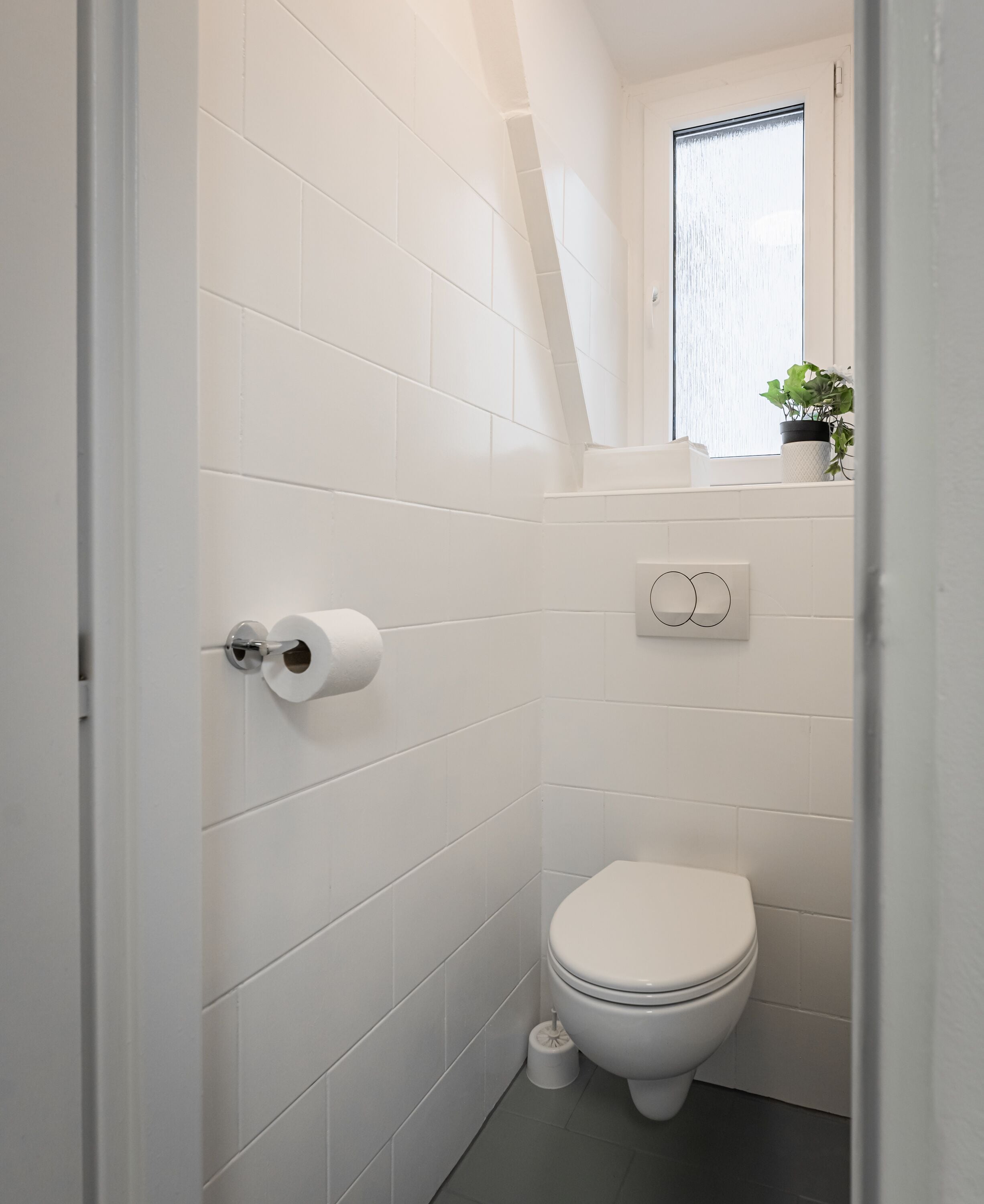 A small, white-tiled bathroom with a toilet, a toilet paper holder, and a window with a plant on the windowsill.