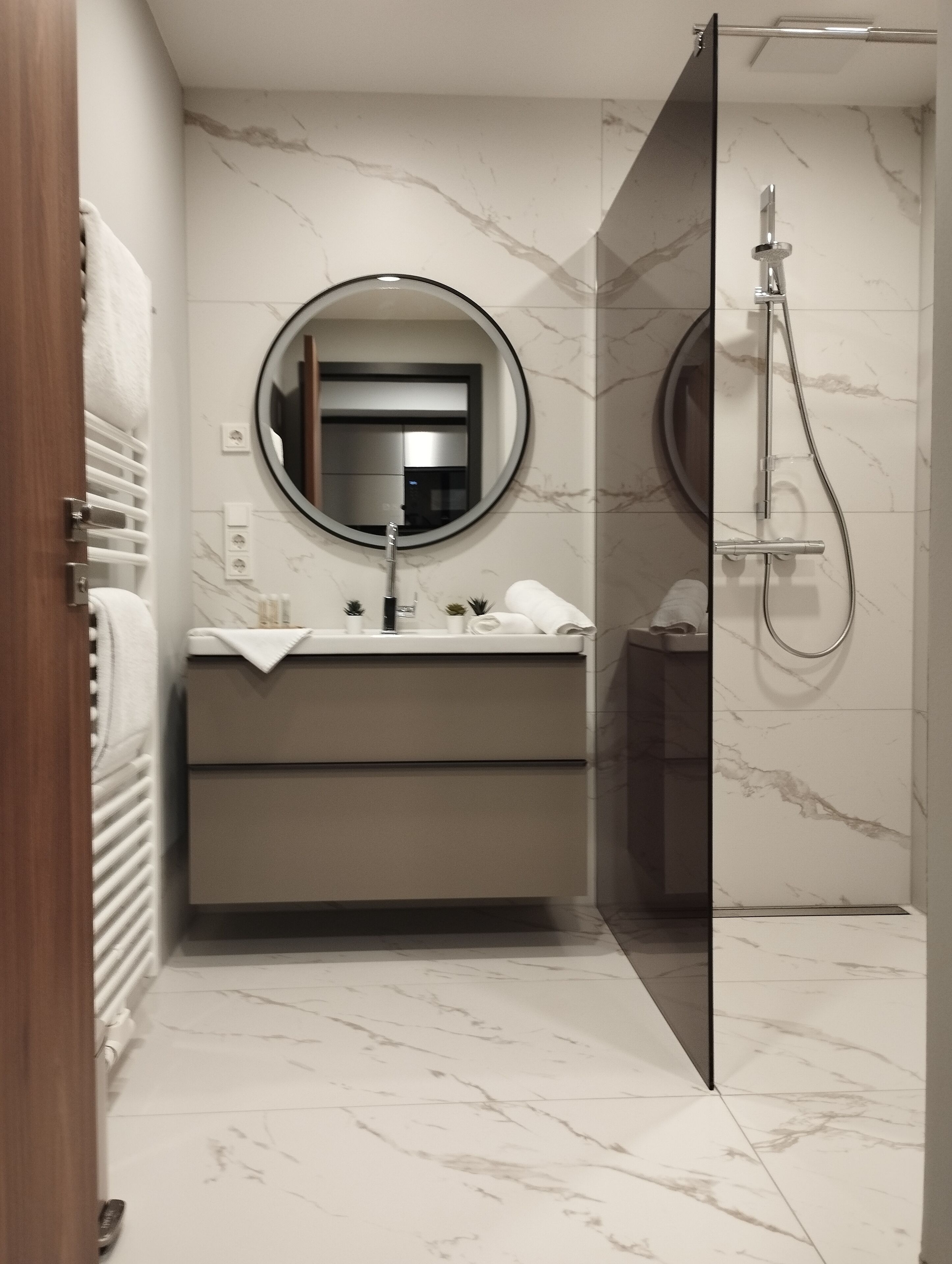 Une salle de bain moderne avec un sol en marbre, un miroir rond, un lavabo avec une serviette et un espace douche.