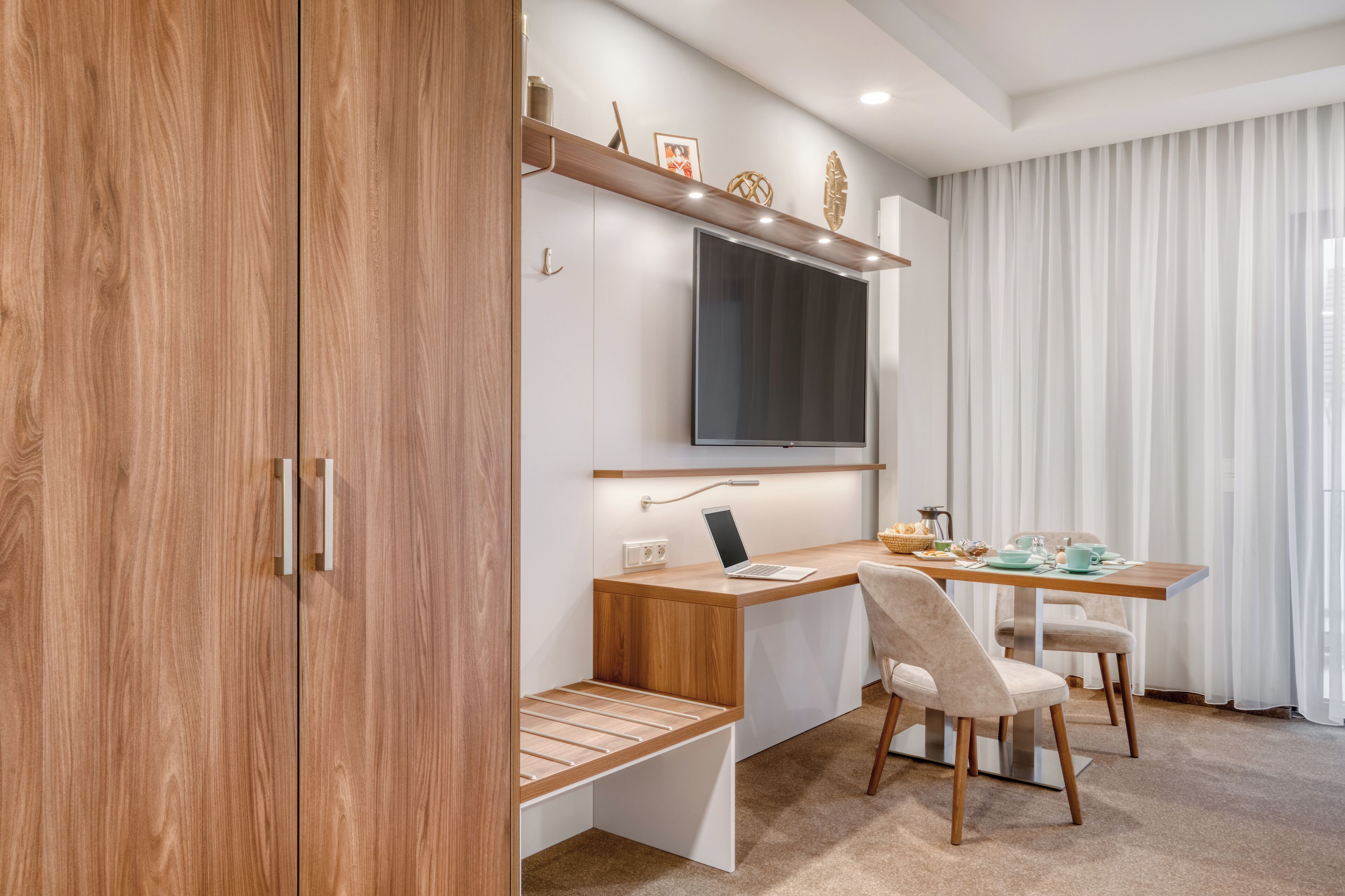A modern kitchen with a wooden cabinet, a built-in desk, a flat-screen TV, and a dining area with chairs and a table.