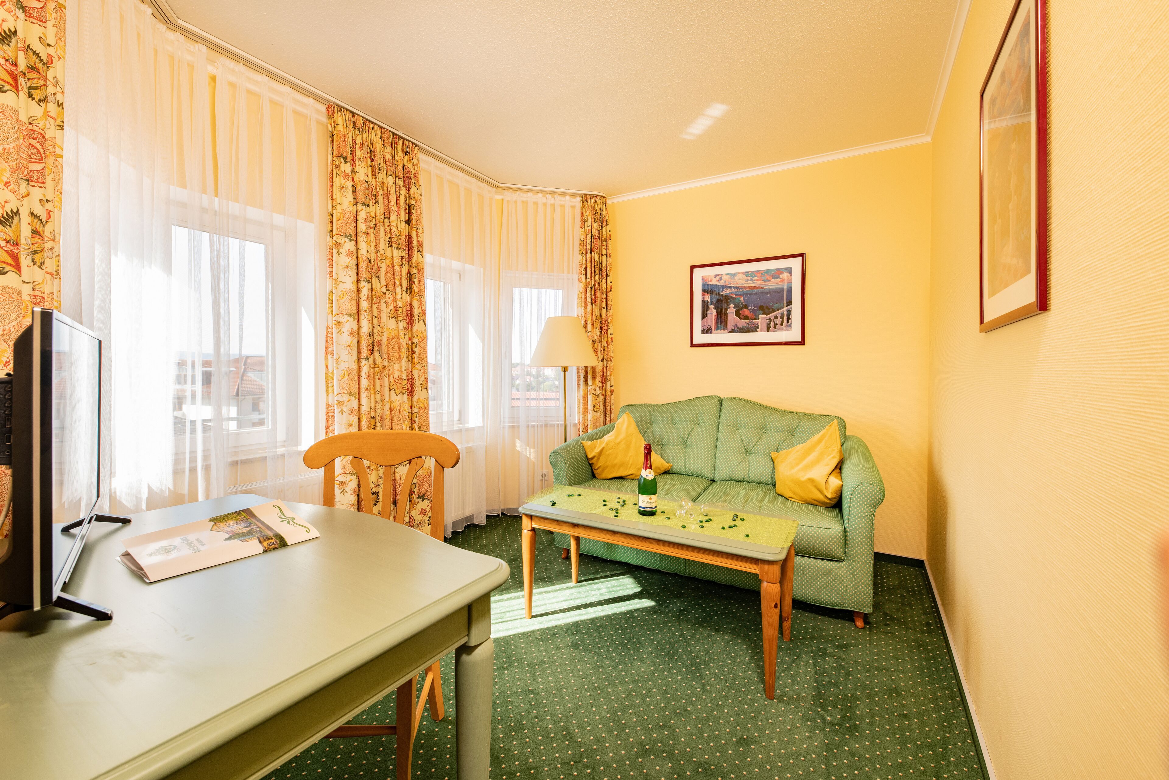 Een hotelkamer met een groene bank, een houten tafel, een lamp en een ingelijst schilderij aan de muur.