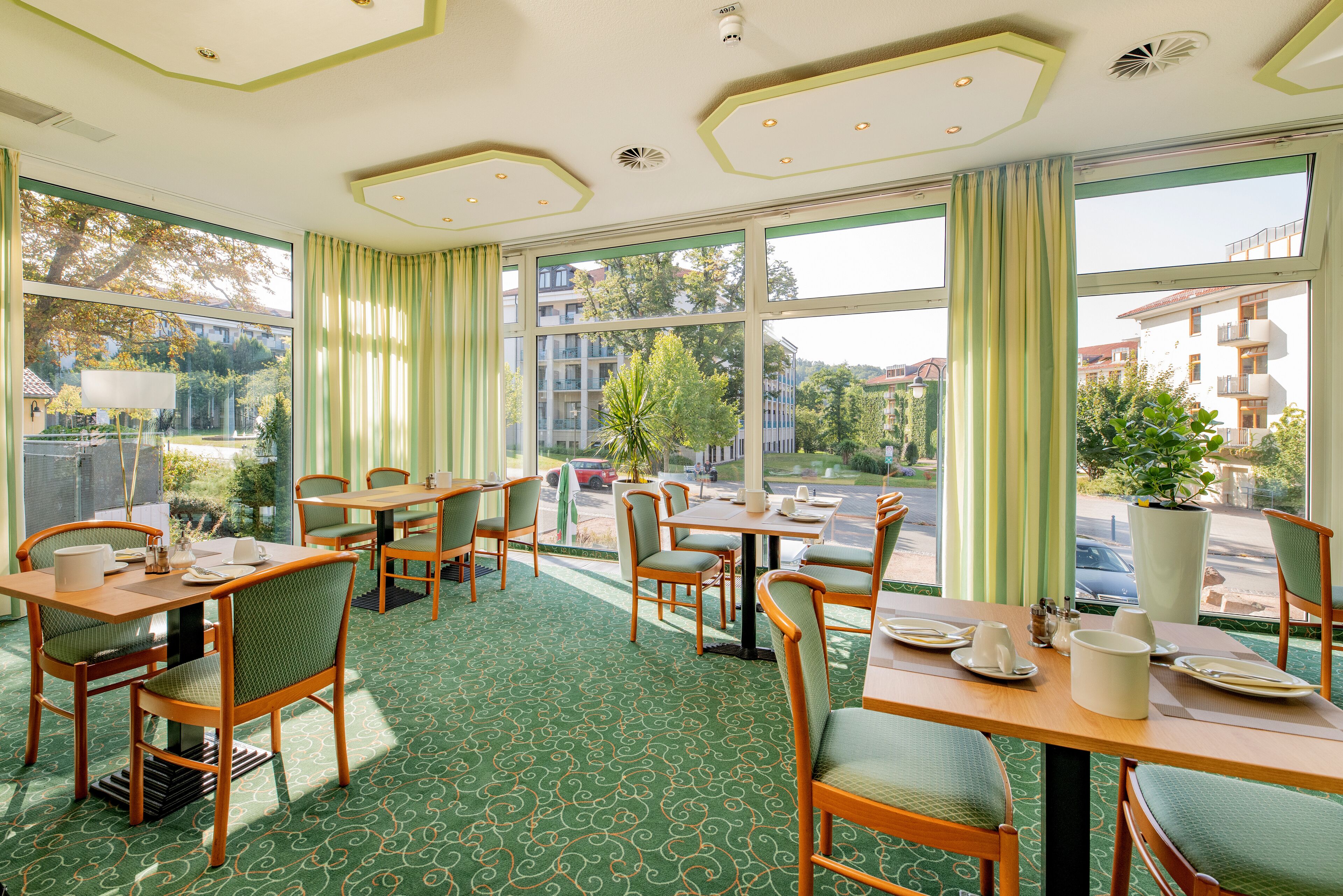 Un comedor de hotel con mesas y sillas, ventanas amplias con cortinas y vistas al exterior.