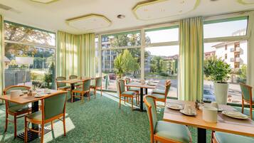 Un comedor de hotel con mesas y sillas, ventanas amplias con cortinas y vistas al exterior.