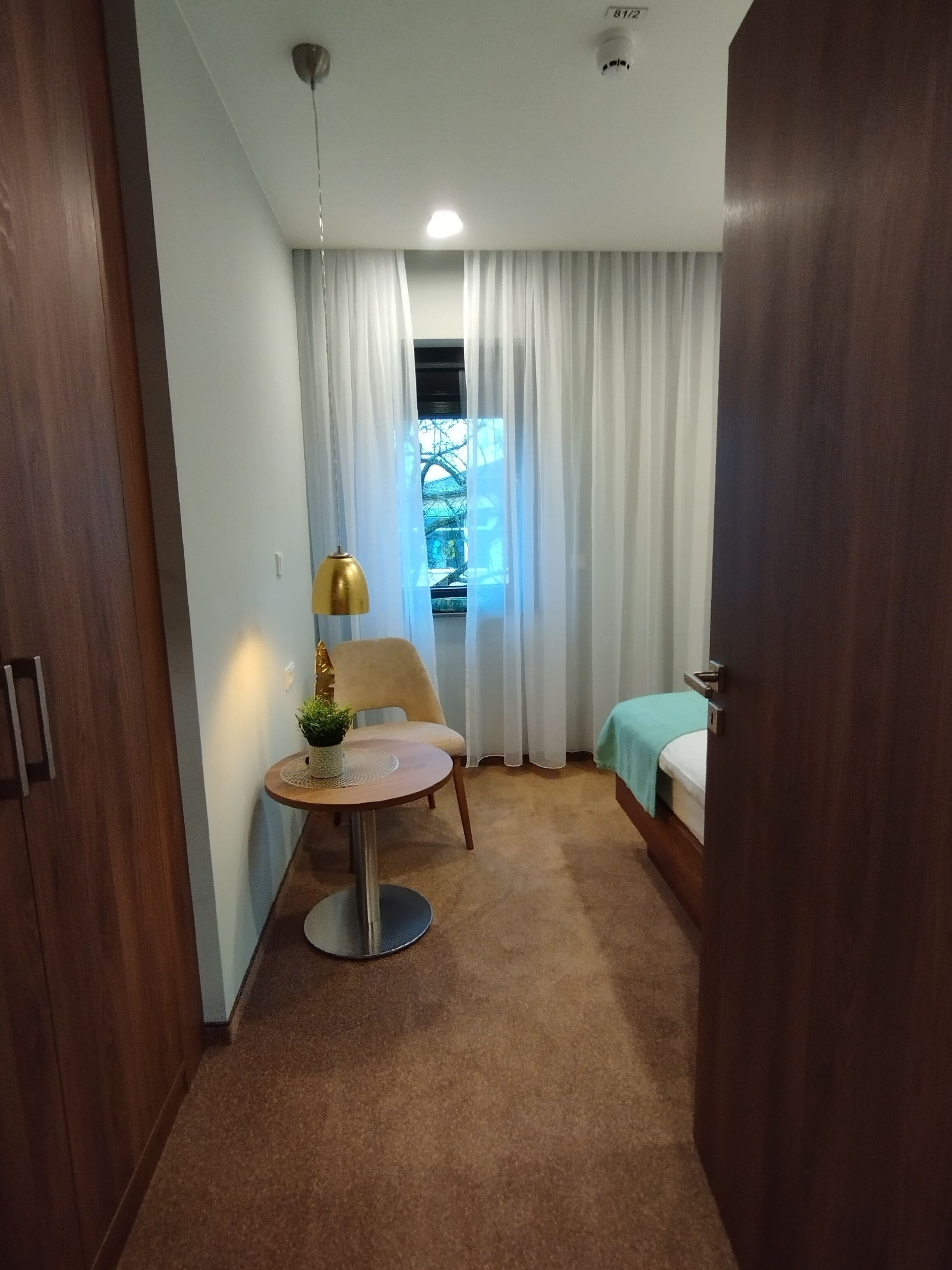 A compact hotel room with a single bed, a small round table with a lamp, a chair, and a window with curtains.