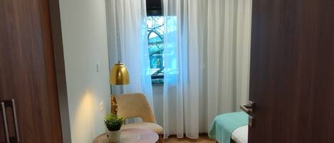 A compact hotel room with a single bed, a small round table with a lamp, a chair, and a window with curtains.