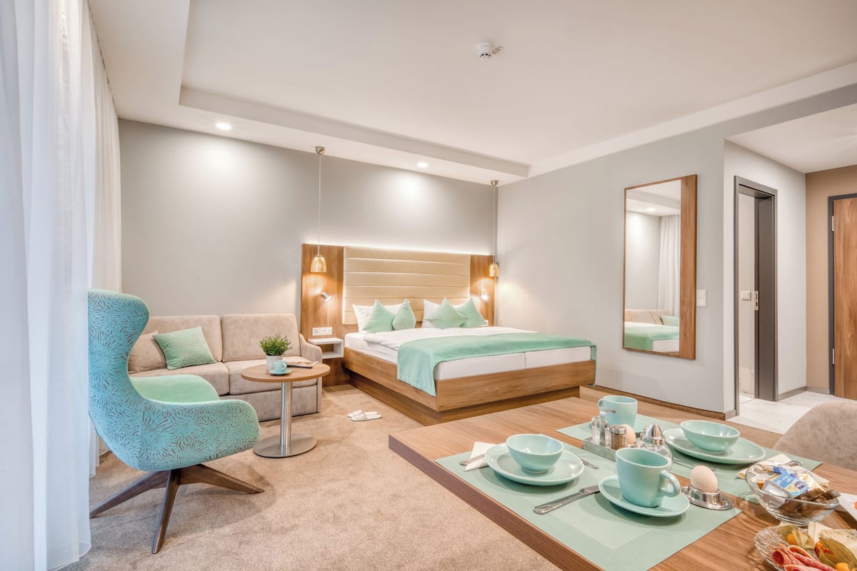 Una habitación de hotel moderna con una cama grande, un sofá, una mesa de comedor con desayuno servido y un espejo amplio.