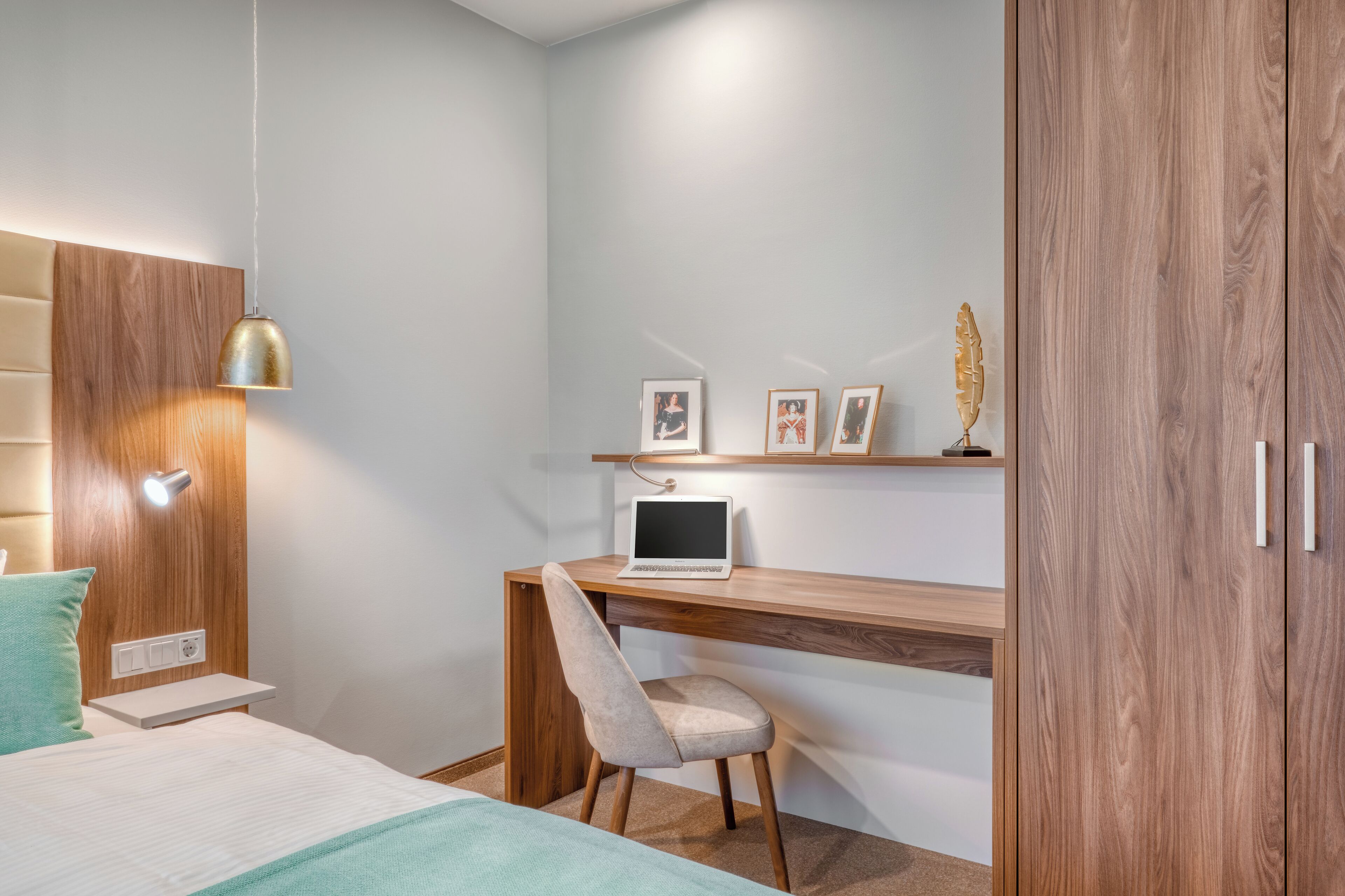 Een slaapkamer met een bed, een bureau met een laptop, een stoel, een plank met foto’s en een kledingkast.