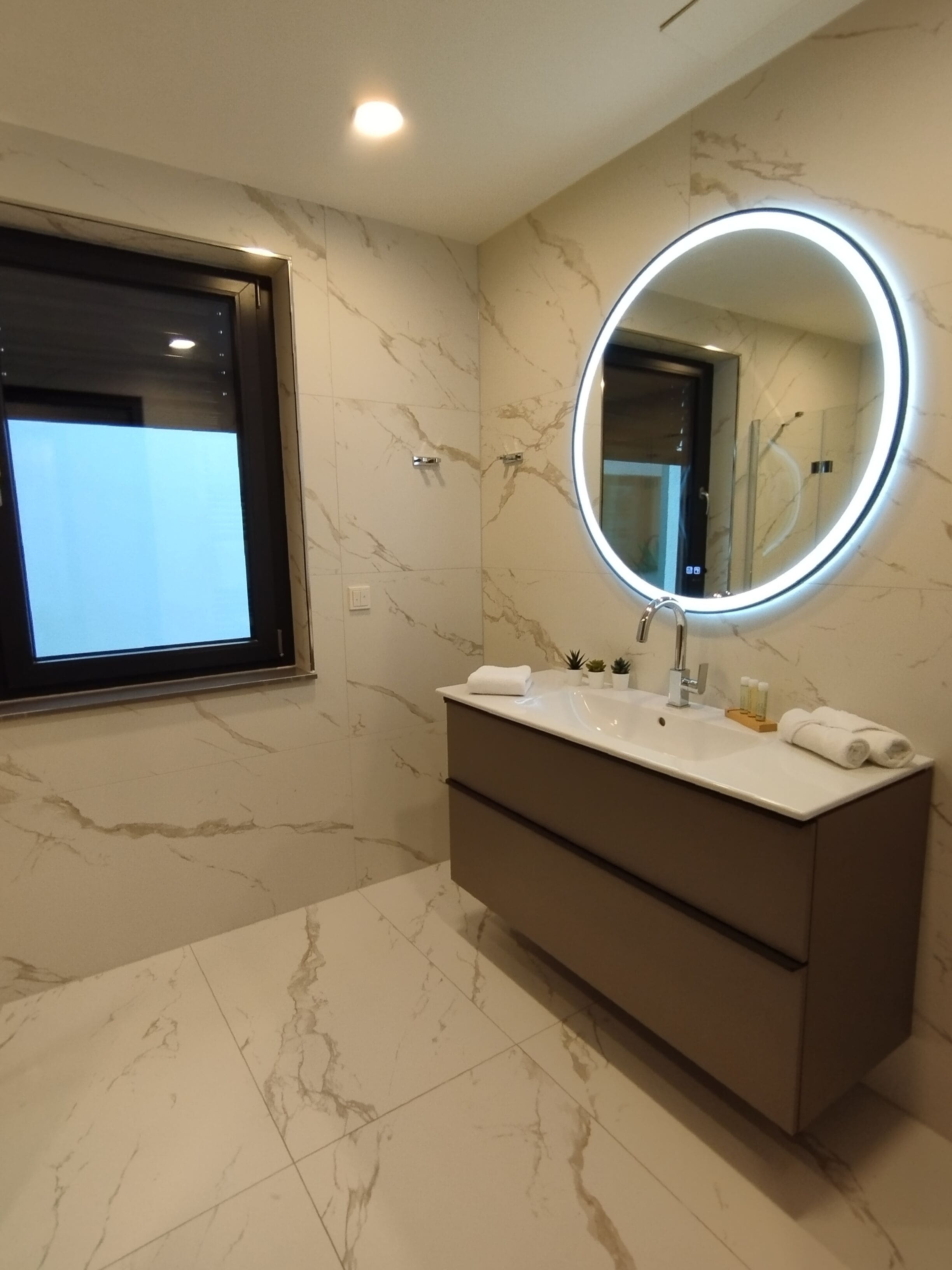Un baño moderno con suelo de mármol, un espejo grande con marco iluminado, un mueble de tocador con lavabo y una ventana sobre el espejo.