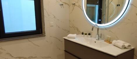 Un baño moderno con suelo de mármol, un espejo grande con marco iluminado, un mueble de tocador con lavabo y una ventana sobre el espejo.