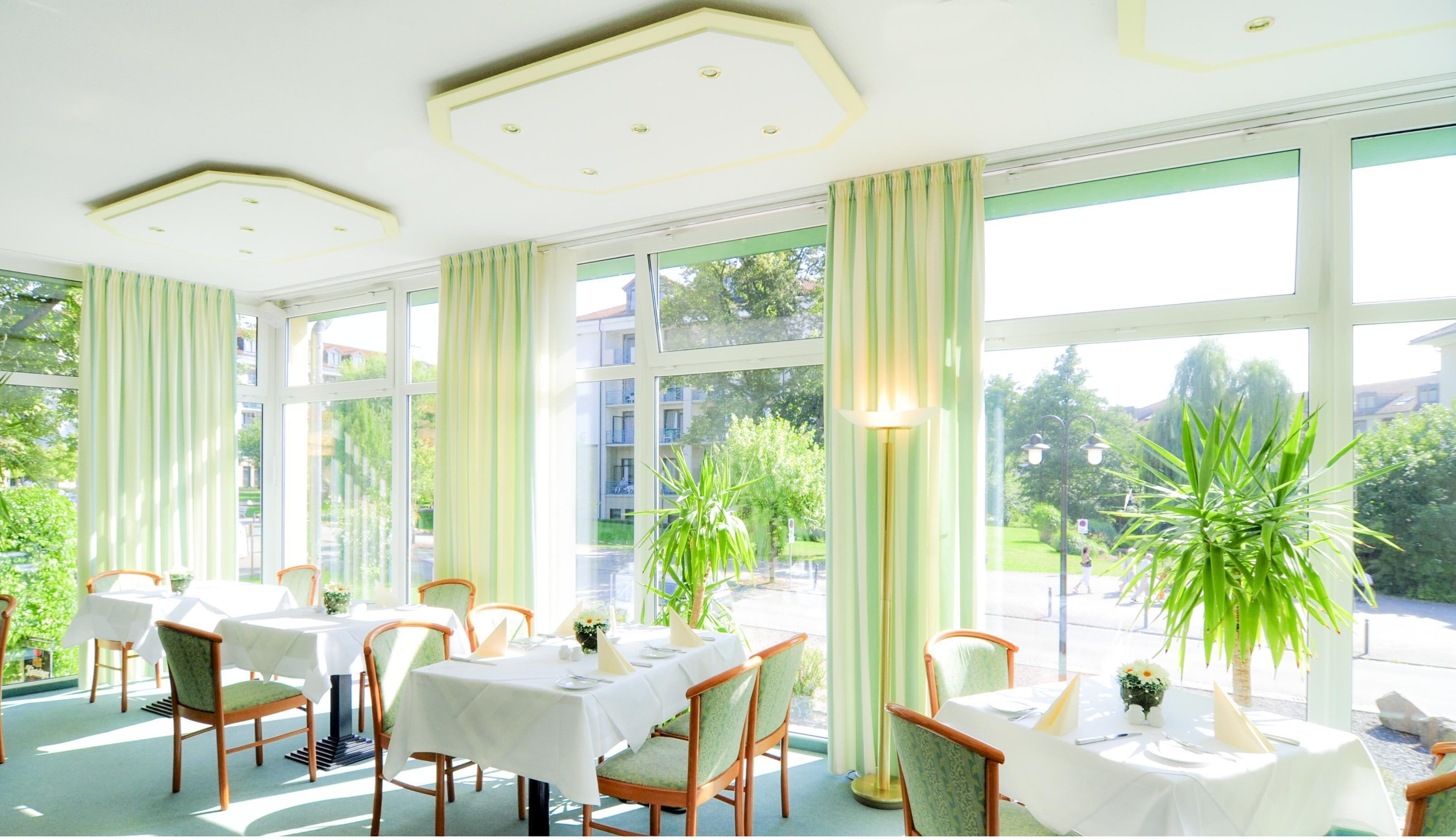 A dining area with tables set for four, large windows, and potted plants.