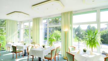 A dining area with tables set for four, large windows, and potted plants.