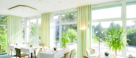 Un comedor con mesas dispuestas para cuatro personas, ventanas amplias y plantas en macetas.