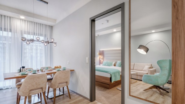 Una habitación de hotel moderna con una mesa de comedor de madera, dos sillas, una cama con cabecera, una lámpara de noche y un espejo grande.