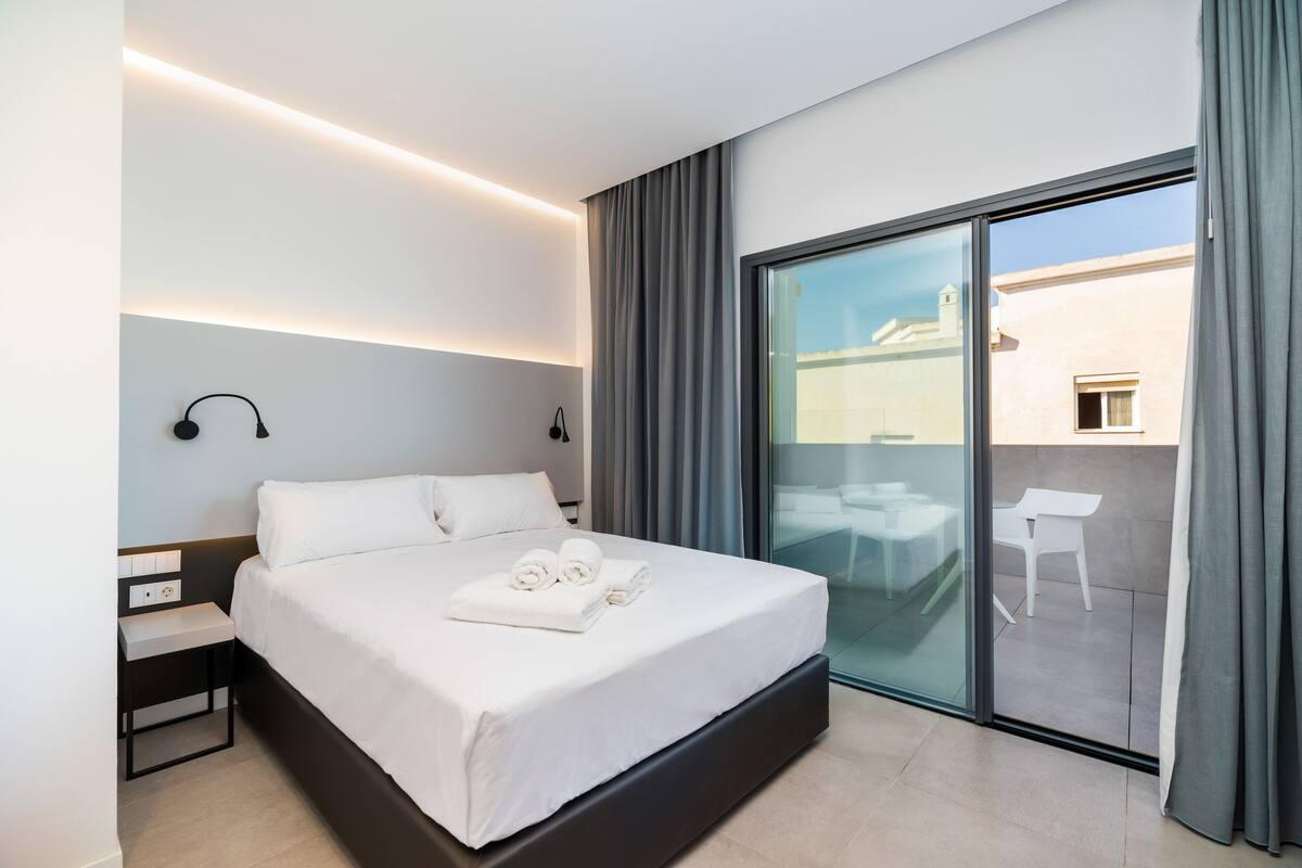 A modern hotel room with a large bed, a bedside table, a wall-mounted lamp, and a sliding glass door leading to an outdoor seating area.