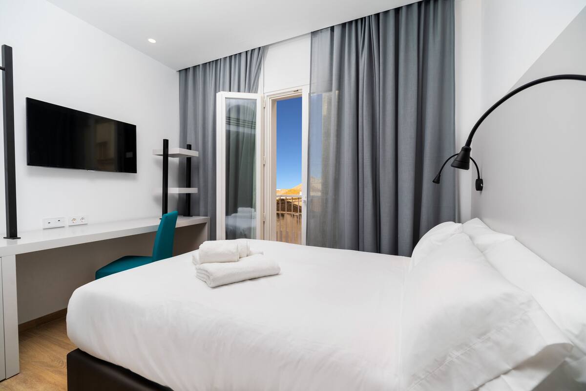 A modern hotel room with a large bed, a desk, a flat-screen TV, and a balcony with curtains.