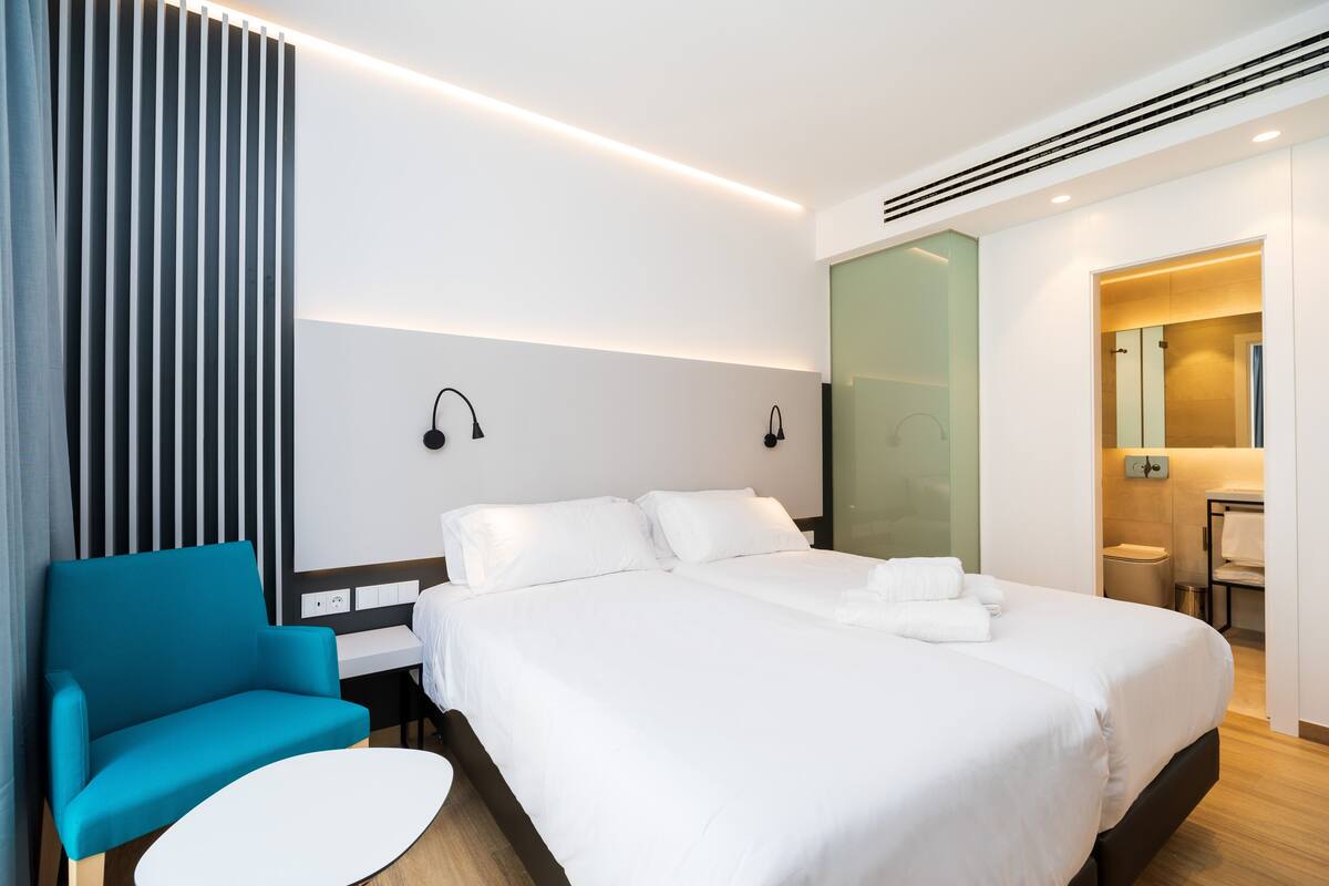A modern hotel room with a large bed, a blue chair, a small round table, and a bathroom visible through an open door.