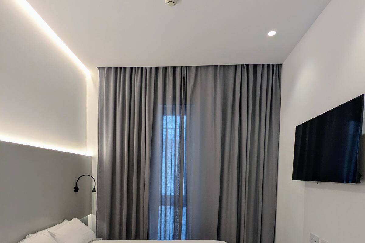 A modern bedroom with a large bed, grey curtains, a mounted TV, and a ceiling light.