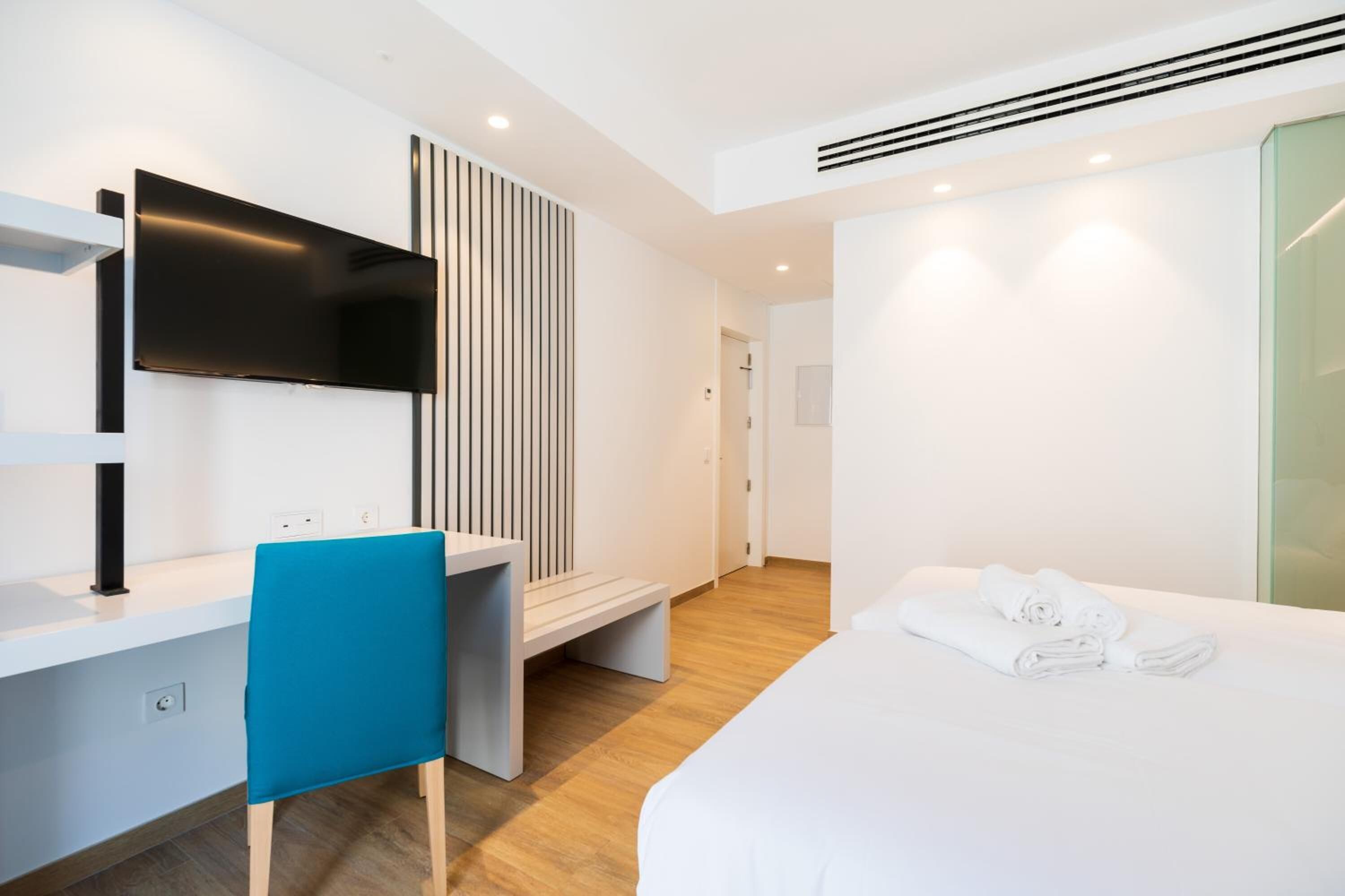 A modern hotel room with a flat-screen TV mounted on the wall, a desk with a blue chair, a bed with white linens, and a glass shower enclosure.