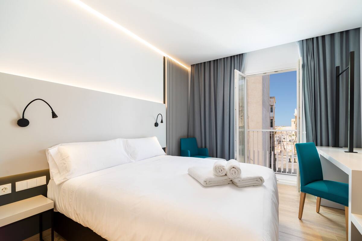 A hotel room with a bed, a desk, a chair, and a balcony with curtains.